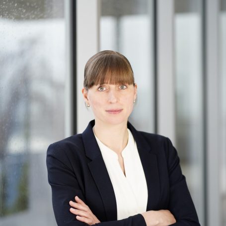 Eine Frau mit blonden Haaren in formeller Kleidung lächelt vor großen Fenstern.